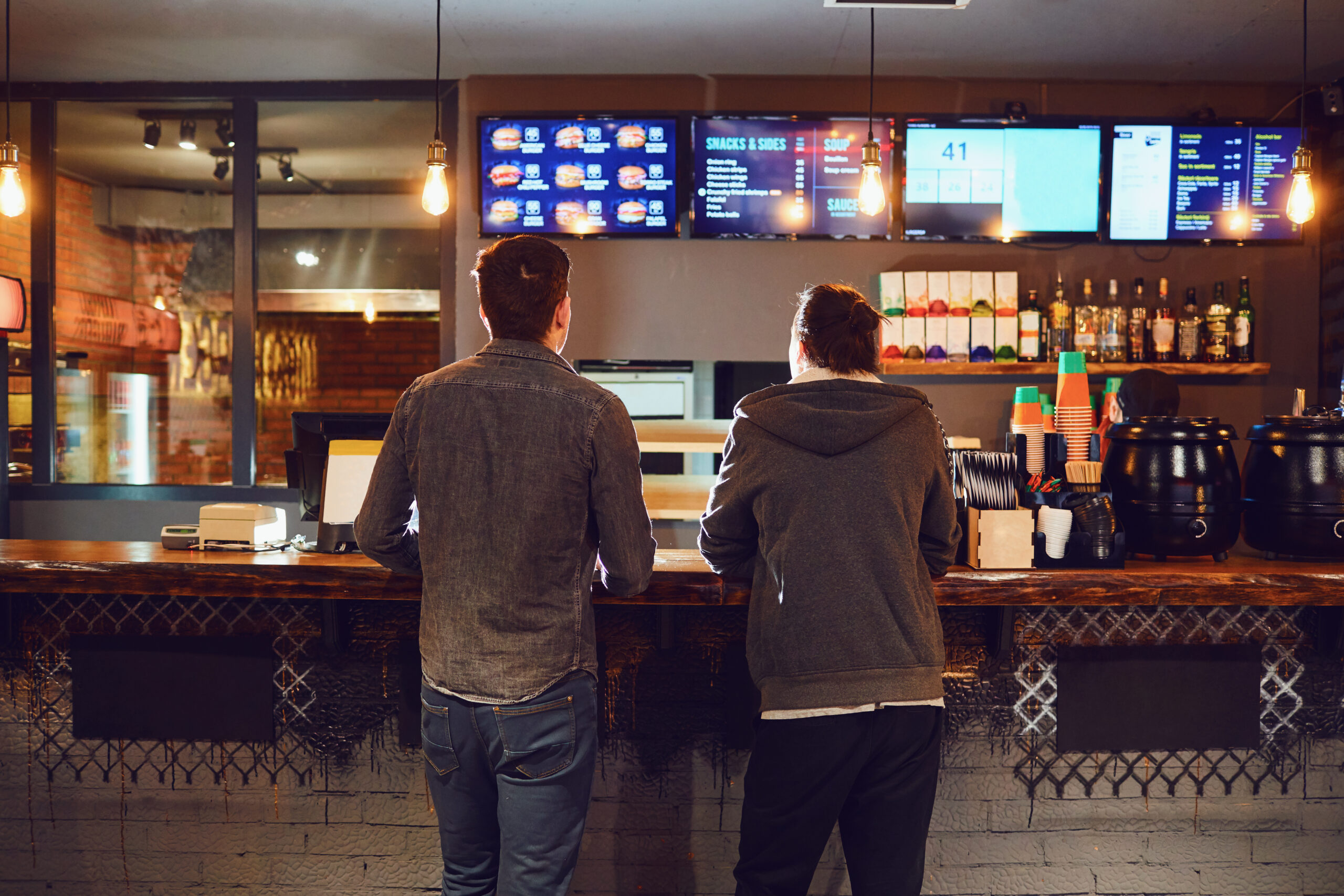 Rear view. Two men choose food in a fast food restaurant looking at a menu determining affordability of options including value meals. By Studio Romantic