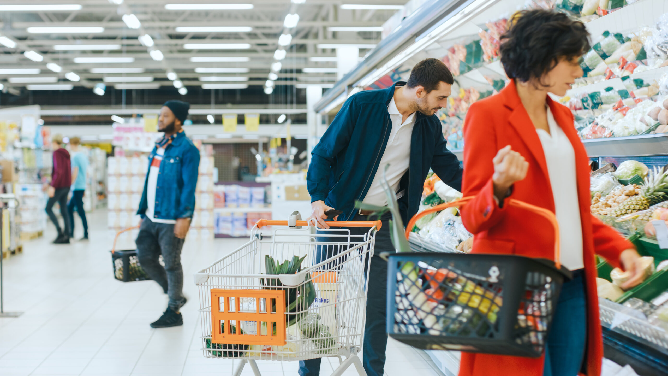 At the Supermarket: Handsome Man with Smartphone, Pushes Shopping Cart, Walks Through Fresh Produce Section of the Store, Chooses Some Products. Other Customers Purchasing Products. 
By Gorodenkoff